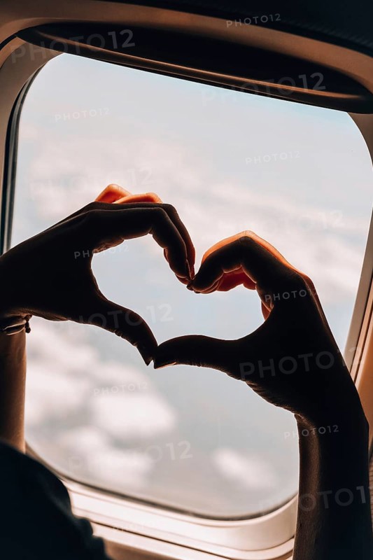 Female hand in heart shape Airplane flight. View from the window of the plane. Airplane, Aircraft. Vacation love travel concept Traveling by air. Airplane Window View Above the Clouds Amazing golden fluffy clouds moving softly on the sky aerial view