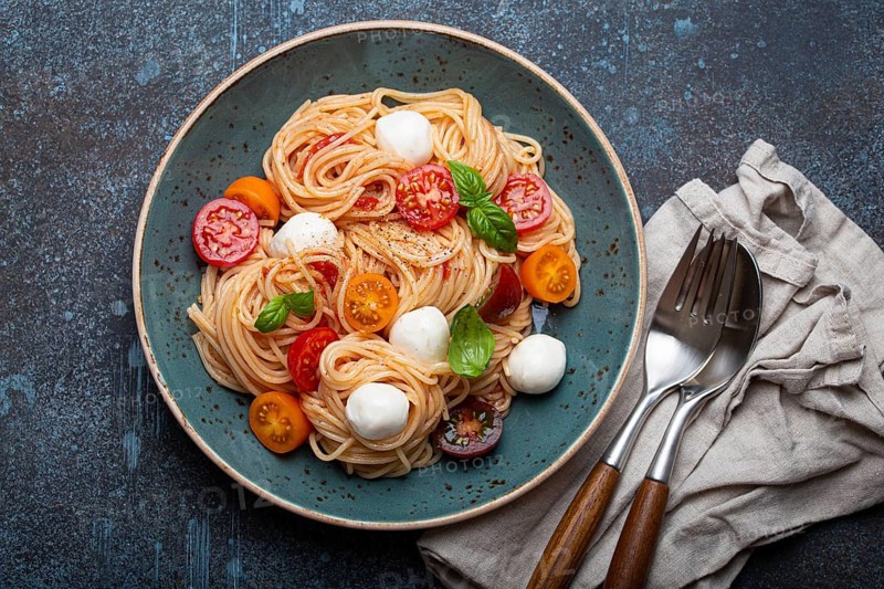 Delicious spaghetti with mozzarella, colourful cherry tomatoes, fresh basil on ceramic plate on rustic stone background top view. Italian healthy tasty food pasta for dinner or lunch, food photography