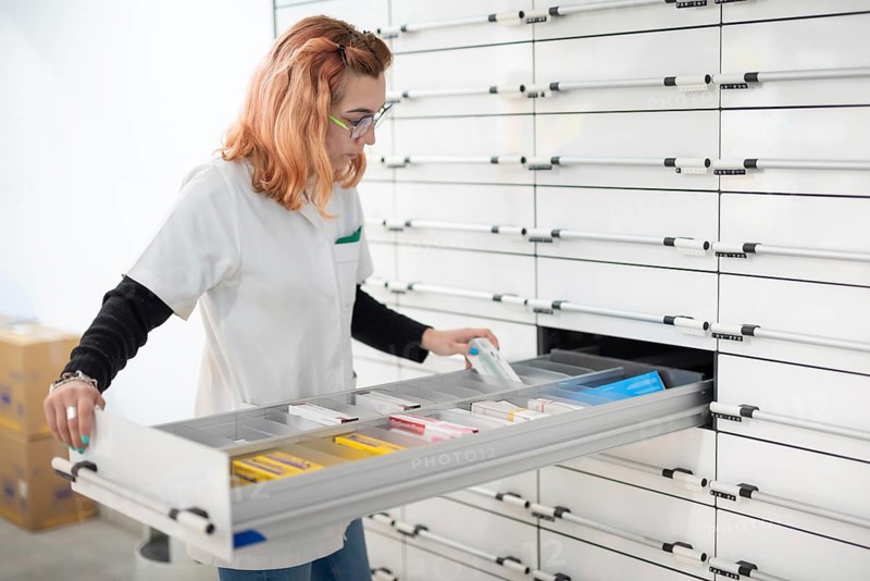 Blonde, straight-haired pharmacist girl with reading glasses, looking for a medicine in the stock of the pharmacy in a drawer of the dispensary