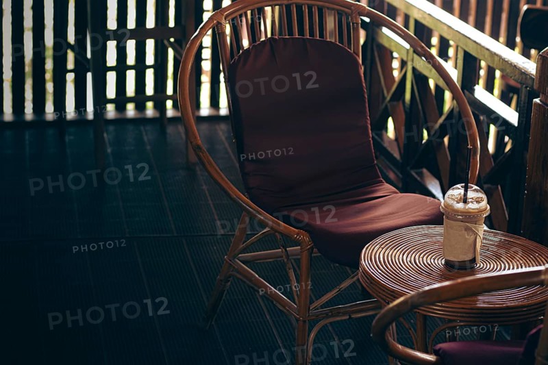 Rattan chair with a brown cushion beside a small circle table holding an iced coffee on a cozy balcony