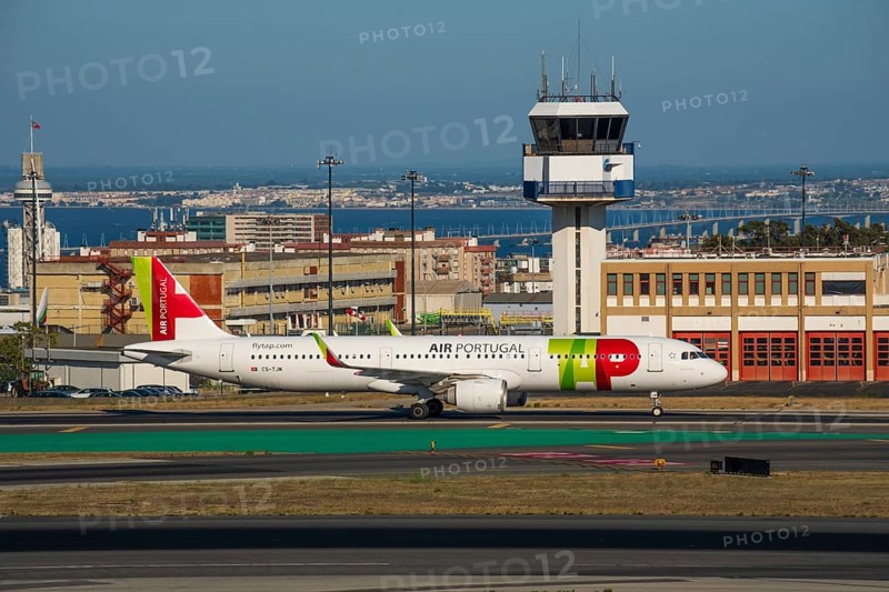 Jul 23, 2023, Lisbon, Portugal. Air Portugal or TAP airplane on runway of Humberto Delgado airport, known as Lisbon airport, in front of ATC tower on sunny day. Residential area and river Tagus in background