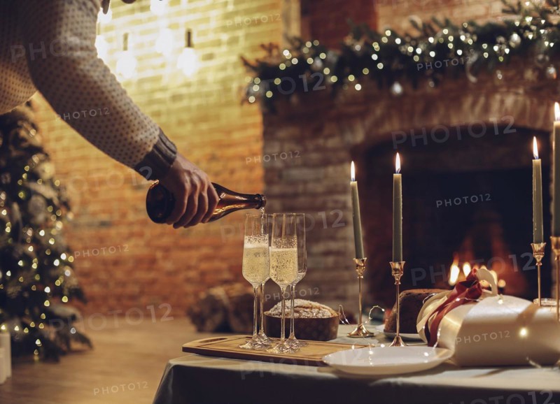 New Years Eve celebration. Man pouring champagne into glasses standing on table with festive xmas dinner, candles and wrapped gifts against blurred background of brick fireplace and firtree, cropped