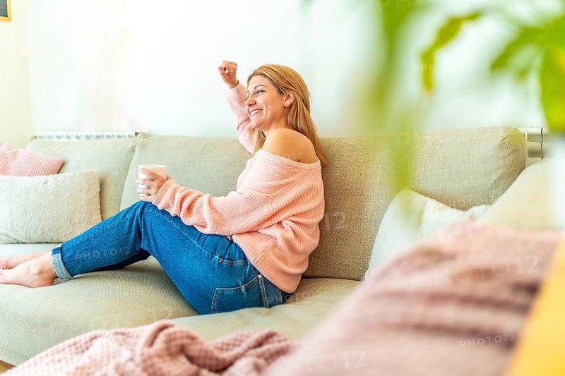 Horizontal photo full length and copy space of a blonde casual adult woman celebrating raising fist sitting relaxed on the sofa