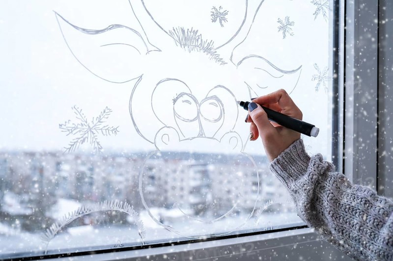 Female hand painting Christmas deer painted on window glass. Christmas winter decor on the window glass. White pattern of deer. Snow art tracery window on holiday season. Diy. Hygge home