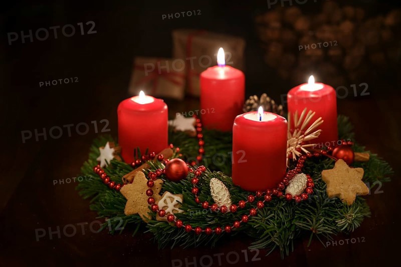 Advent wreath from fir branches with red lit candles and Christmas decoration, some gifts blurred in the dark background, copy space, selected focus, narrow depth of field
