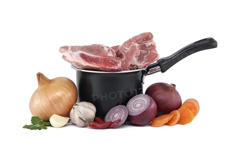 Black saucepan with pieces of raw pork inside. Around the saucepan are sliced vegetables which include onions, carrots, and garlic isolated on white background. Preparation of meat broth
