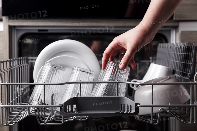 Close-up of female hand loading dished to, empty out or unloading from open automatic stainless built-in dishwasher machine with clean utensils inside in modern home kitchen. Household domestic life