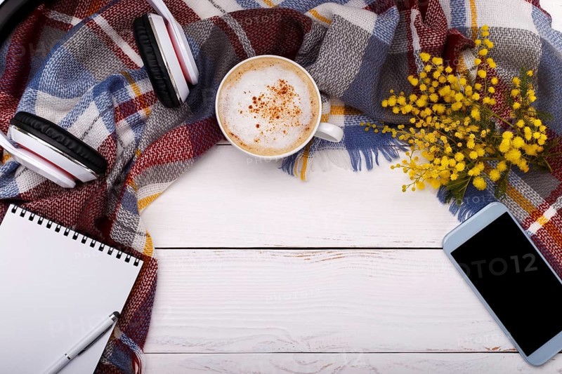 Hot coffee cappucino, blanket, mimosa flowers, notepad, smartphone and head phones on old wooden background. Seasonal relax concept, flat lay