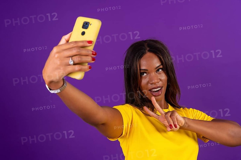 Studio portrait with purple background of an african urban woman gesturing success while taking a selfie with mobile phone