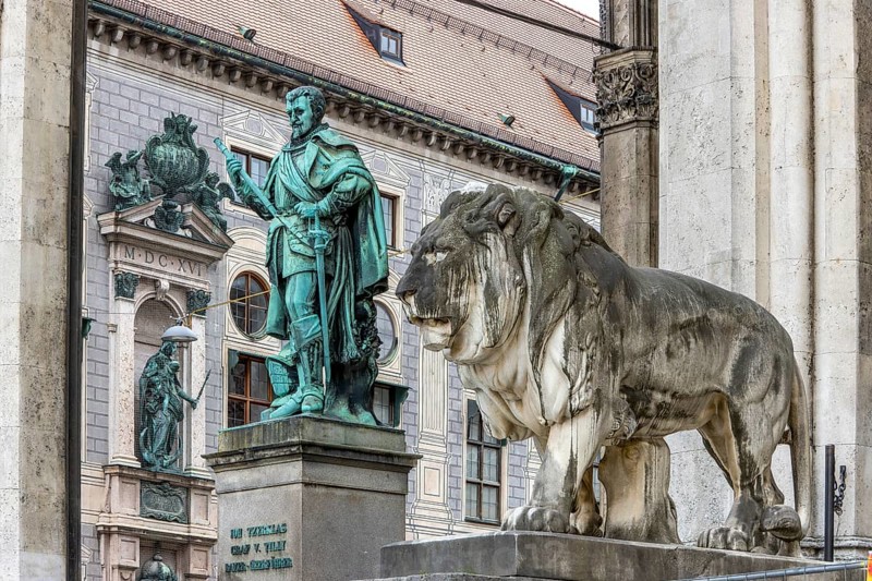 Stone sculpture lion, bronze sculpture by Johann Tzerklas Count of Tilly in front of Feldherrnhalle and in the background the sculpture on the building of the Munich Residence, Odeonsplatz, city centre, Munich, Upper Bavaria, Bavaria, Germany, Europe