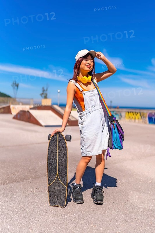 Asian woman holding a skateboard and adjusting her cap standing on a skateboard park