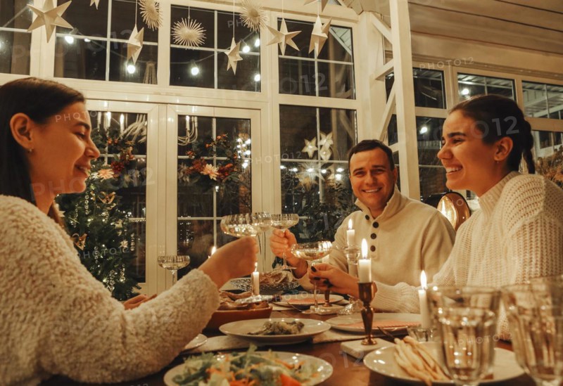 Happy family couple clinking champagne glasses while celebrating New Years Eve or Christmas with friends, sitting at festive dinner table with traditional food and burning candles