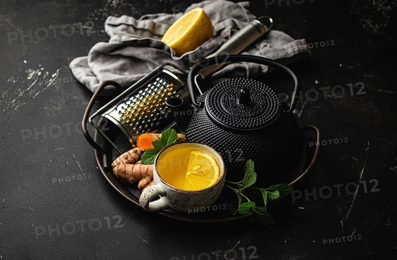 Healthy warm herbal turmeric tea for boosting immune system. Ingredients for making detox tea in cup, black teapot, fresh curcuma root, lemon, mint leaves, grater on black stone background