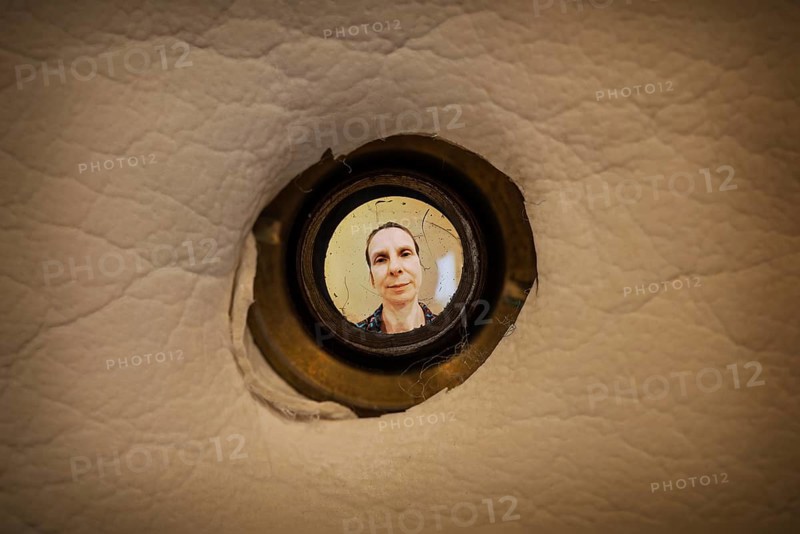 Woman seen out of focus through the old dirty peephole of the front door of a dark apartment
