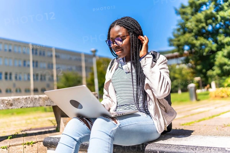 African female student with braided long hair using laptop outside the campus in a sunny day