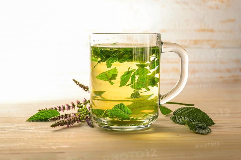 Fresh peppermint leaves brewed as a healthy herbal tea in a glass cup, some flowering stems on a light wood table, copy space, selected focus, narrow depth of field