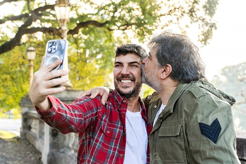 A man is kissing his husband while his husband takes a selfie in that scene