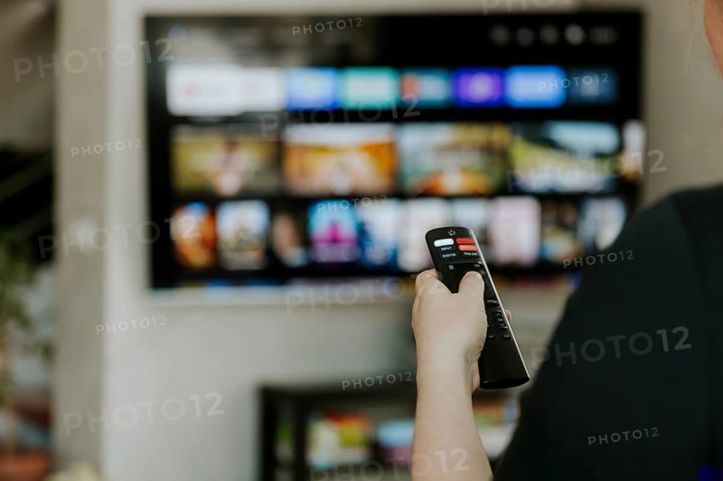Hand holding remote control pointing to TV. Person watching smart television indoors. Blurred background. Colorful screen, copy space. Woman finger in foreground pressing button. Movie stream service