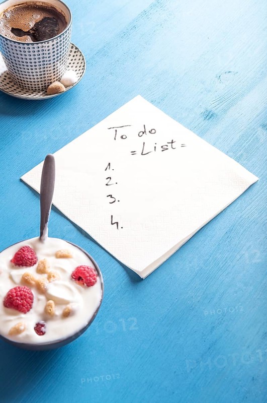 White napkin with a draft of a to do list, surrounded by a bowl of yogurt with cereal and raspberry and a mug of aromatic coffee, on a blue background
