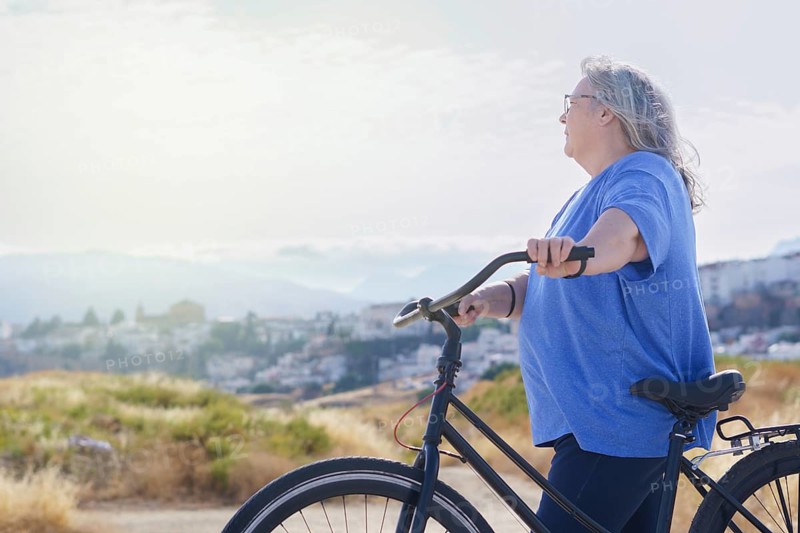 An overweight mature woman rides a black retro-style bicycle against a cloudy mountain backdrop with a village in the background