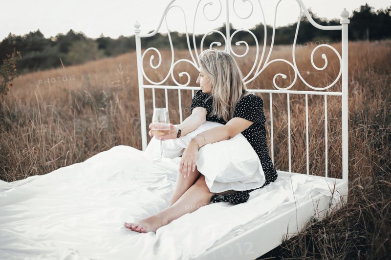 Full body barefoot female with blond hair enjoying wine and looking away while sitting on comfortable bed in grassy field in nature