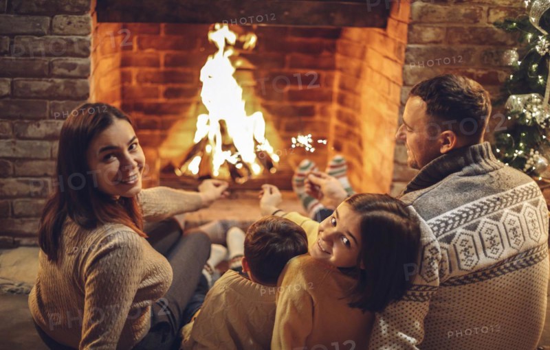 Happy young family parents with two small kids gathering around brick fireplace in cozy and warm country house during Christmas holidays in winter. Father, mother ad children sitting by fire at home