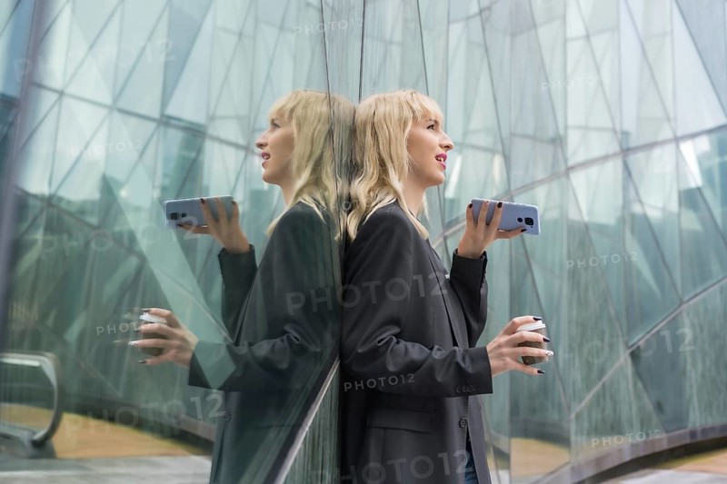 Entrepreneurial girl business park in a modern building reflected in the windows, young blonde in a suit sending a voice message with a take away coffee