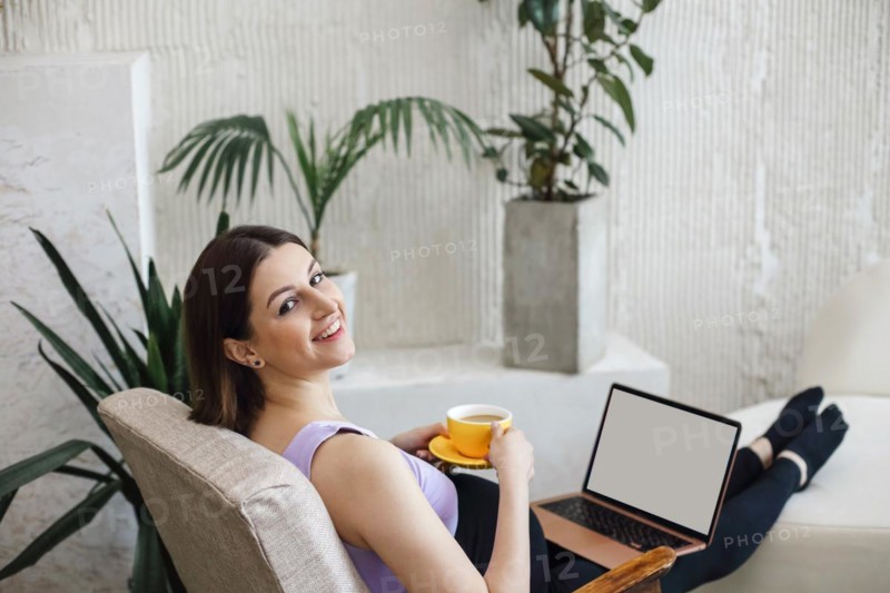 Happy pregnant young adult woman sitting on a sofa, working on a laptop and holding a cup of coffee. Pregnancy at home concept