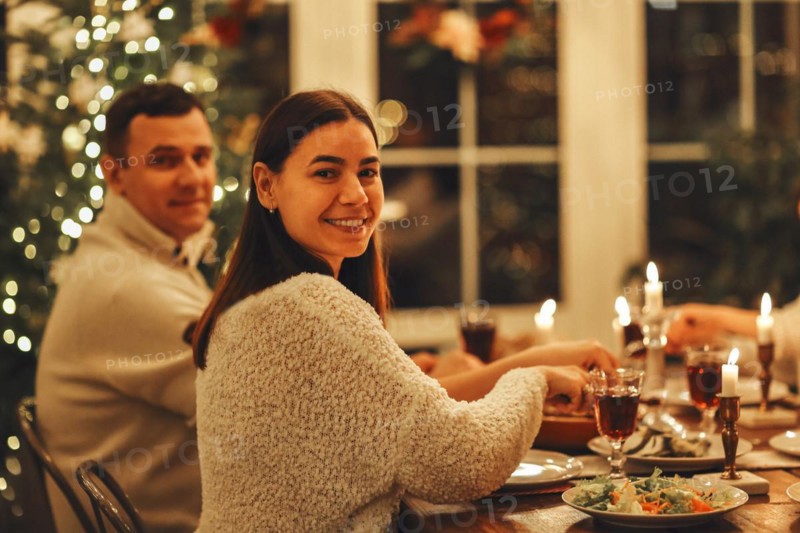 Happy young family couple smiling at camera while sitting at festive dinner table served for Christmas eve, husband and wife celebrating New Year at cozy decorated home with xms tree in background