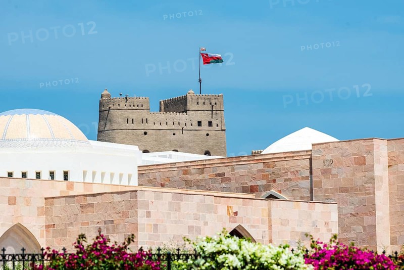 An old fortress with the Omani flag rises above pink stone buildings under a blue sky, Al Alam Palace and Al Jalali Fort, Muscat, Oman, Asia