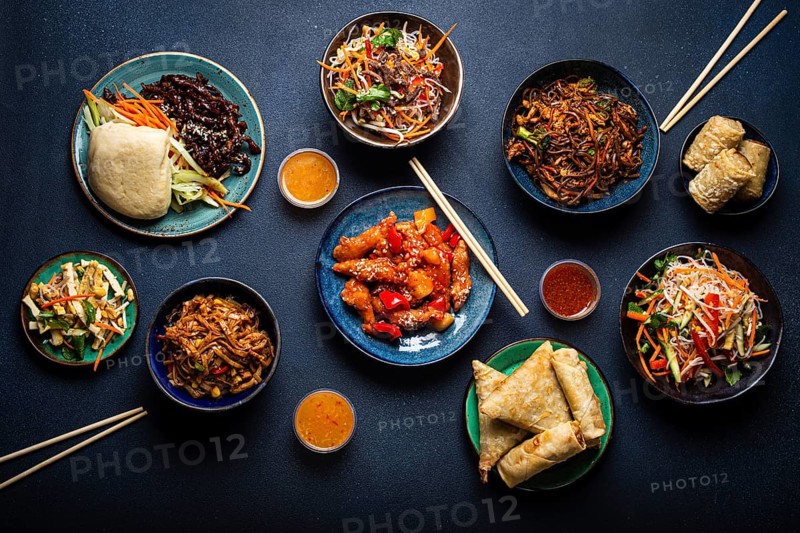 Set of assorted Chinese dishes on table: sweet and sour chicken on plate, fried spring rolls, noodles, salad, rice, steamed buns with bbq glazed pork, Asian style banquet or buffet, top view