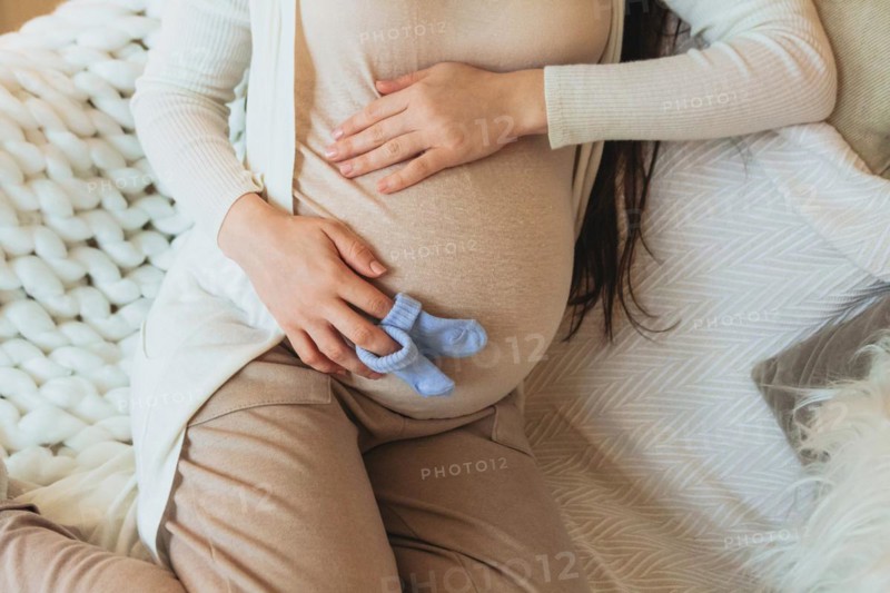 Maternity and pregnancy concept. Cropped photo of pregnant woman holding pair of cute blue baby socks for unborn child on belly while relaxing at home. 9 months in anticipation of baby