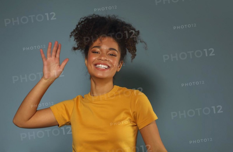 Beautiful happy dark skinned african american ethnicity woman in yellow tshirt raising hand in greeting while looking in camera with pleasant smile, posing against gray green studio wall background