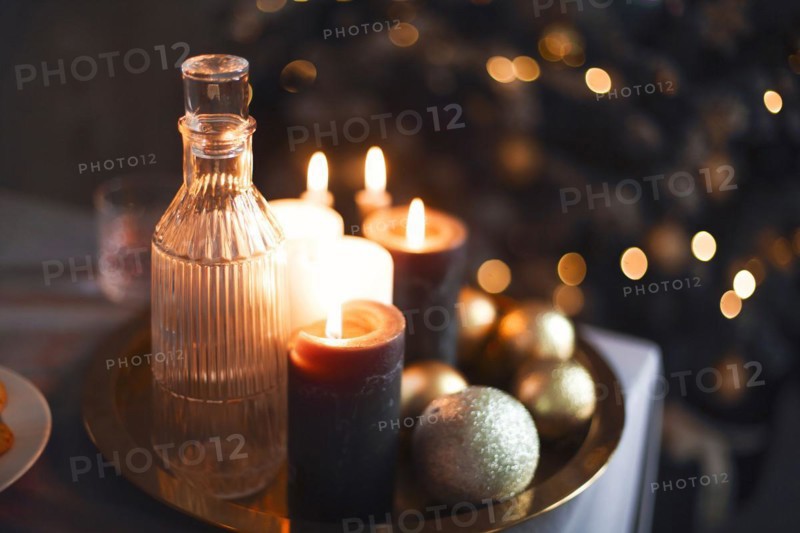 New Year's still life. Glasses and a decanter from transparent glass, candles and golden xmas balls. Festive feast. The table is laid by a black cloth. The burning candles ad Christmas tree background