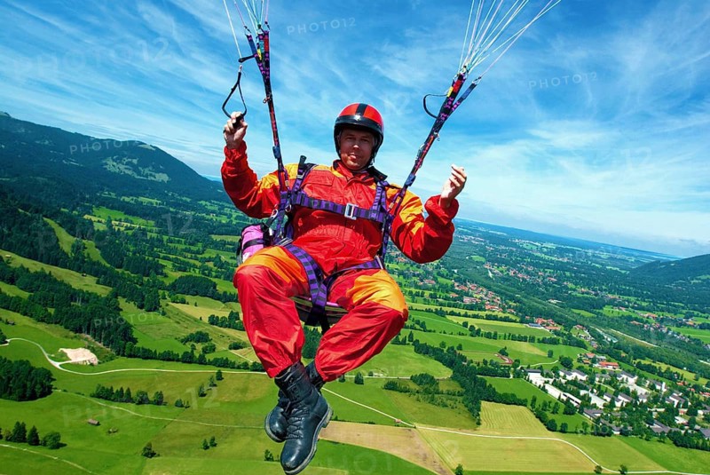 A paraglider in orange and red overall flies over the meadows at Brauneck near Lenggries, Bavaria, Germany, Europe