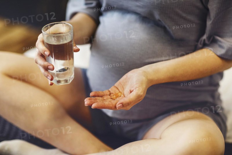 Supplements during pregnancy. Young happy pregnant woman taking prenatal vitamins, holding glass of water and pill while sitting on bed at home. Expectant mother beginning each day with folic acid