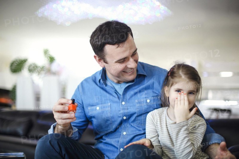 Cheerful father playing with little daughter sitting on couch and helping to paint her nails with bright polish play together. Young brunette father with his cute little daughter at home