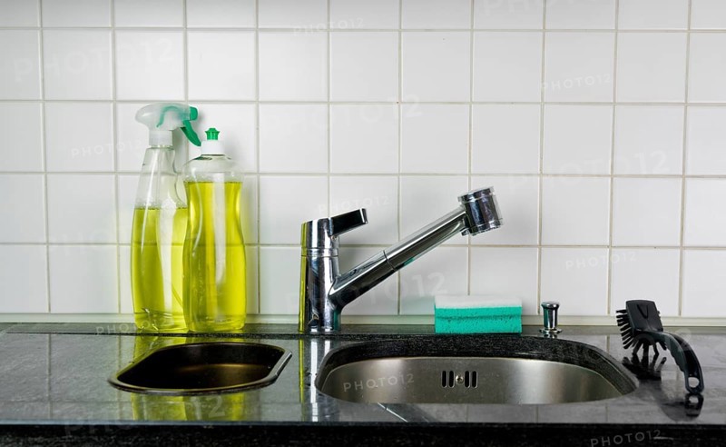 kitchen sink and faucet with a sponge and brush on a black granite table top and yellow kitchen cleaner and dishwashing liquid behind