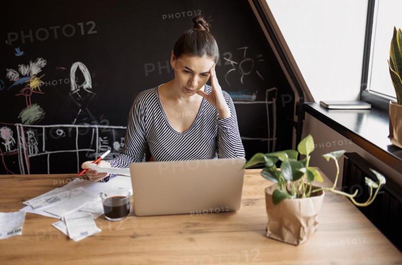 Serious housewife sits at table at home kitchen check household bills, make calculations on calculator, pay utilities through e-banking system on laptop. Manage family finances concept