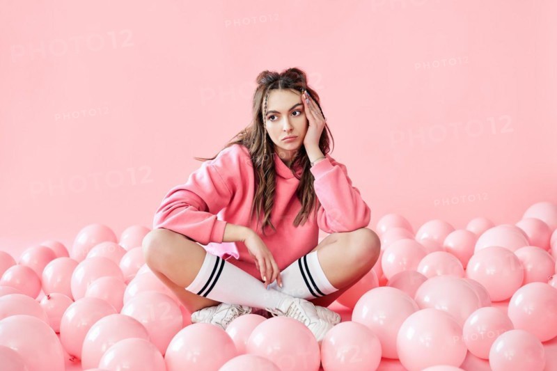 Bored fashionable woman sitting on floor with pink air balloons on pink pastel background. Celebration, bad party, disappointment concept
