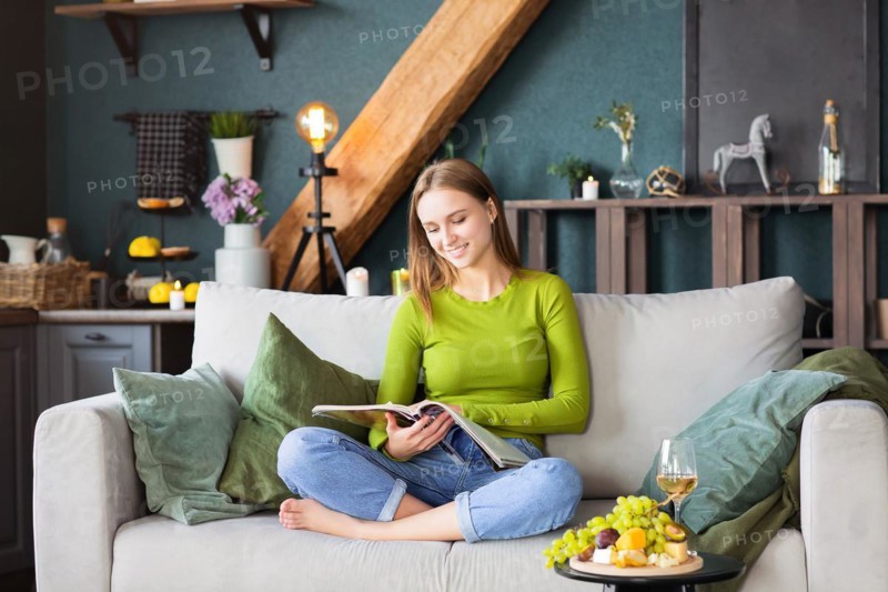 Young blond female reading interesting magazine while sitting on comfortable couch near table with fruits and wine on weekend day at home