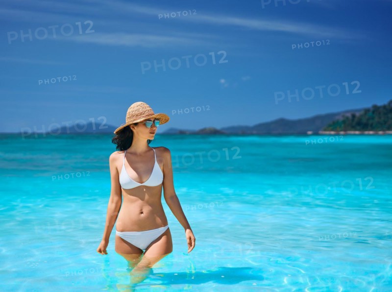 Sexy young woman in straw hat and white bikini on tropical paradise beach. Summer vacation, travel destination, relax concept