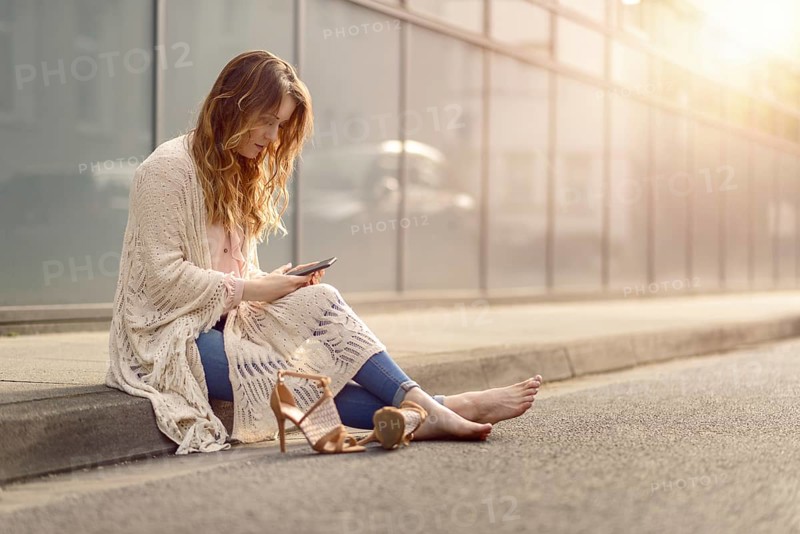 Attractive trendy woman sitting on the kerb of an urban street with her high heeled shoes lying on the ground alongside her as she reads an sms on her mobile phone, with copy space