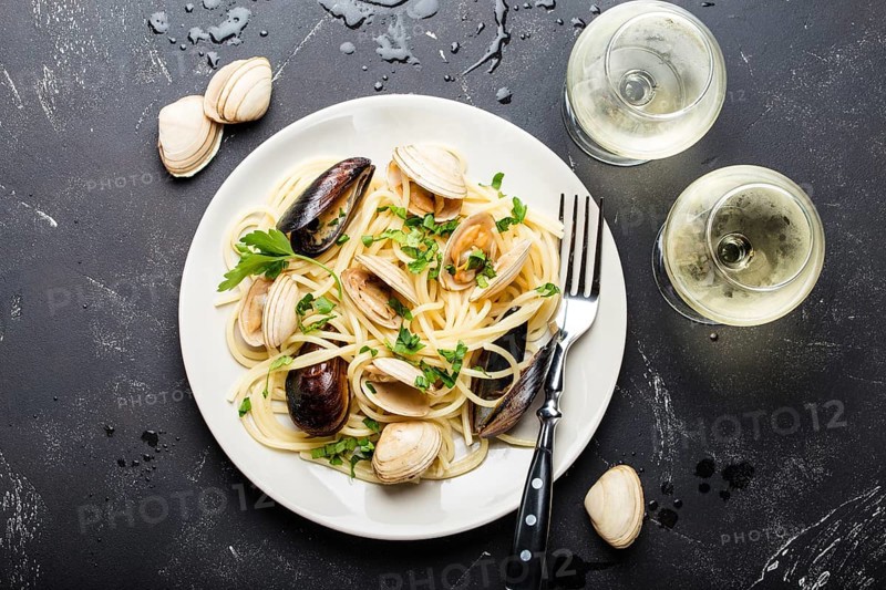 Spaghetti vongole, Italian seafood pasta with clams and mussels, in plate with herbs and two glasses of white wine on rustic stone background. Traditional Italian sea cuisine, close-up, top view