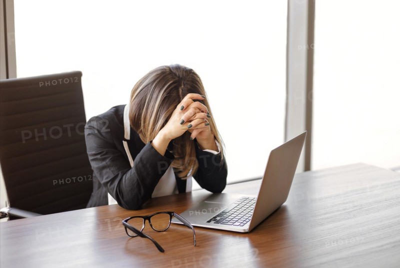 Young female manager taked of glasses after typing on netbook keyboard while sitting at table and working in modern office. Tired employee, emotional burnout concept