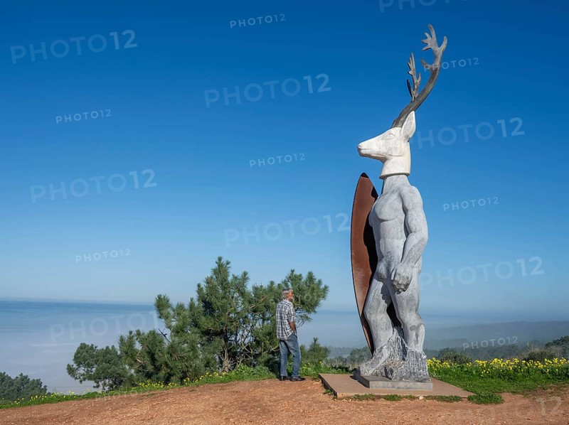 A man stands in front of a surfer statue with a deer head, Nazaré, Portugal, Europe