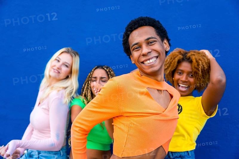 Close-up of a transgender person dancing happily with multi-ethnic friends next to a blue wall