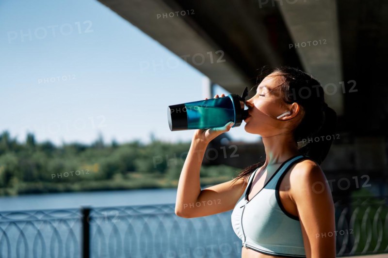 Young fitness woman drinking water from bottle. Sporty female resting after work out exercising in industrial city background near river