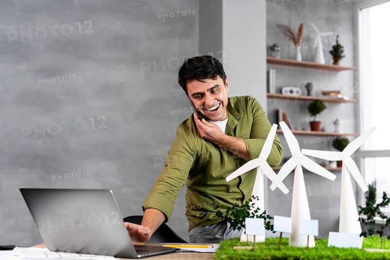 Smiley man working eco friendly wind power project while talking phone using laptop