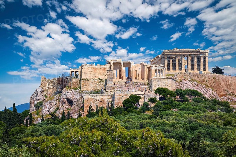 View of Acropolis hill with Propylaea and Parthenon in Athens, Greece from Pnyx hill in summer daylight with great clouds in blue sky. Great fresh foliage of olives and cedar trees in foreground. Close shot. Awesome composition